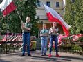 Harcerze ubrani w mundury harcerskie stoją na scenie trzymając flagi Polski. Scena znajduje się na dwrzoe w tle widać drzewa i budynek szkoły 