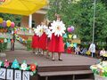 Uczennice z nauczycielkami trzymając kwiatki tanczą na scenie na dworze. Scena jest udekorowana kwiatami i balonami. W tle widać drzewa i bud