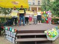 Uczniowie stoją na scenie na dworze trzymając etykiety z napisami miłość poświęcenie opieka wychowanie troska cierpliwość. Scena jest udek