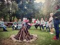 Harcerze ubrani w mundury harcerskie siedzą na ławce w otoczeniu zieleni przed nimi znaduje się przygotowane drewno na ognisko poznają obrządek rozpalania harcerskiego ogniska
