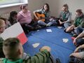Harcerze z druhnami ubraniw mundury harcerskie siedzą w kręgu na dywanie jedna osoba trzyma flagę Polski jedna druhna gitarę śpiewają pieśni patriotyczne