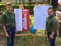 Dwóch harcerzy ubranych w mundury harcerskie stoją przed sztalugami z plakatami w tle widać zieleń huśtawkę głośnik budynek