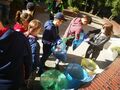 Uczniowie znajdują się na terenie przed szkołą trzymają worki ze śmieciami segregują śmieci do worków odpowiedniego koloru powieszone przed nimi na stojakach