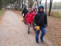 Harcerze ubrani w kurtki z kapturami na głowie idą scieżką w otoczeniu drzew trzymajac w rękach liściew oddali podąża grupa ludzi