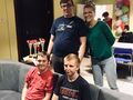 Dwóch uczniów siedzi na kanapie. Za nimi stoi uśmiechnięty naucyciel obok uczeń w granatowej koszulce. Na stole puchary