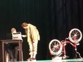 Teatralna scena. Pierwszy chłopiec siedzi przy biurku drugi pochyla się nad nim. Obok czerwony rower 