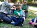 Druhna zaklada innej drugnie temblak wykorzystując do tego chustę harcerską. Pozostali harcerze siedzą dookoła nich na trawie i przygladają się