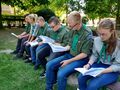 Harcerze ubrani w mundury harcerskie siedzą na ławce trzymają w rękach śpiewniki 