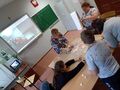 2 Uczniowie z nauczycielem tworzą plakat. Smarują klejem i przyklejają elementy na arkusz szarego papieru