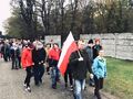 Uczniowie wraz z nauczycielami idą w pochodzie przez miejscowość Konarzew z przodu uczeń trzyma flagę Polski