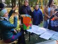 Pani Iza siedzi w parku na krześle w ręku trzyma instrument muzyczny. Dzieci z nauczycielami stoją przed ławką i rozwiązuja zagadkę dźwiękową 