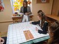 Uczennice i nauczycielka znajdują się dookoła ławki szkolnej na której jest mata do kodowaniawykonują zadanieukładając elementy na macie 