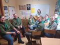 Grupa harcerzy wraz z druhną znajdują się w sali lekcyjnej. Są ubrani w mundury harcerskie druhna trzyma gitarę 