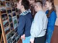 Grupa uczniów stoi przy tablicy znajdującej się na sztaludze w szkolnym holu. Na tablicy znajdują się zdjęcia z przygód harcerskich 