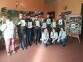 Harcerze wraz z druhnami pozują do zdjęcia w szkolnym holu przy tablicy ze zdjęciami na sztaludze. Harcerze trzymają w rękach kartki z pokolorow