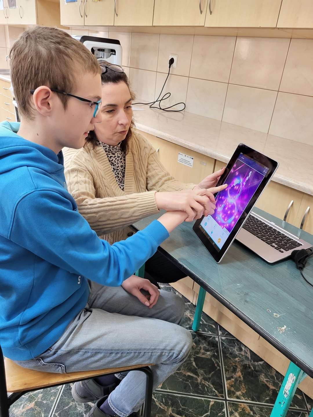 chłopiec siedzi przy stole i rozwiązuje zadanie na tablecie wspomagany przez nauczycielkę