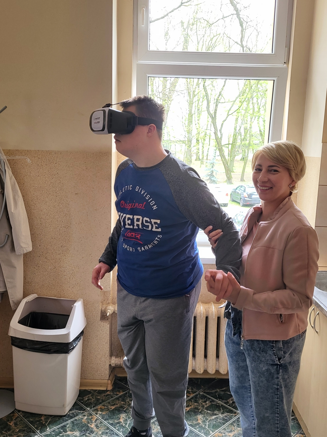chłopiec stoi trzymany za rękę przez nauczycielkę ma na głowie okulary wirtualnej rzeczywistości