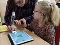 dziewczynka siedzi przy stole i rozwiązuje zadanie na tablecie wspomagana przez nauczycielkę