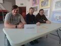 Trzech uczniów siedzi w sali przy ławce na której postawiona jest kartka z napisem szkoły. W tle widać stojące krzesła i wiszące obrazki na ści