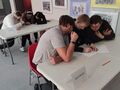Uczniowie siedzą przy ławkach w trzyosobowych drużynach wykonują zadania konkursowe. W tle widać stojące krzesła i obrazki wiszące na ścianie 