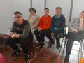 Ucznowie siedzą na krzesłach w muzeum i słuchają pogadanki