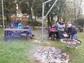 uczniowie siedzą w ławkach przy stołach przed nimi ognisko nauczyciel rozkłada kiełbaski na ruszcie