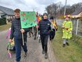 Uczniowie maszerują ulicą. Na pierwszym planie widać uczniów trzymających transparent Witaj Wiosno. W tle widać uczennicę przebraną za Panią Wio