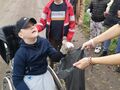 Chłopiec na wózku inwalidzkim wrzuca śmieci do worka. Znajduje się na drodze gruntowej w otoczeniu zieleni 