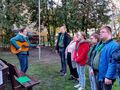 Harcerze stoją w szeregu. Nauczycielka gra na gitarze. Śpiewają pieśni patriotyczne. Znajdują się w parku w tle widać flagę Polski na maszcie 
