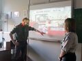 Nauczycielka i uczeń ubrani w mundury harcerskie znajdują się przed tablicą interaktywną uczeń włącza element dźwiękowy w prezentacji interakty