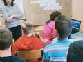 6. Uczennica pracuje przy komputerze, obok stoi nauczycielka, reszta uczniów siedzi na krzesłach