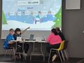 uczniowie siedza przy stole na stole 2 laptopy na scianie wyswietlana prezentacja na planszy