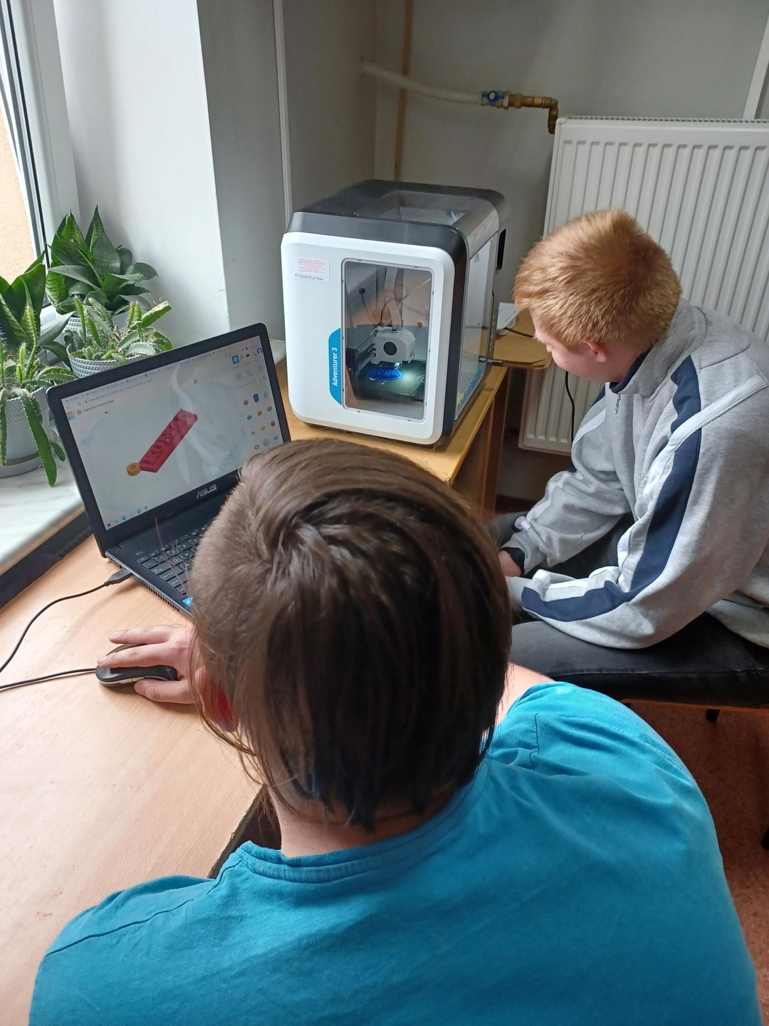 8. Dwóch uczniów siedziprzy ławce jeden obsługuje na laptopie program do tworzenia projektów 3d drugi obserwuje proces drukowania przez drukar