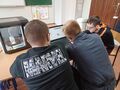 6. Troje uczniów siedzi przy ławce szkolnej obsługują na laptopie program do druku 3d. Obok laptopa znajduje się drukarka 3d w trakcie drukowan