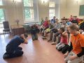 1 Uczniowie i nauczyciele siedzą na krzesłach w klasie przed nimi policjant klęczy na podłodze z rękami za głową