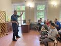 2 Uczniowie i nauczyciele siedzą na krzesłach w klasie przed nimi stoi policjant z uniesioną ręką