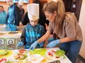 Nauczycielka pokazuje uczniowi w którym miejscu ma położyć warzywo na kanapkę. 