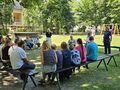 dzieci siedzą na ławkach w parku i słuchają pani policjantki która stoi i mówi