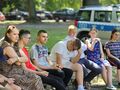 uczniowie i nauczyciele siedzą na ławkach w parku i słuchają pani policjantki która stoi i mówi w tle stoi radiowóz