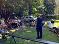 uczniowie siedzą na ławkach w parku i słuchają pani policjantki która stoi i mówi w tle stoi radiowóz
