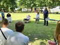 uczniowie siedzą na ławkach w parku i słuchają pani policjantki która stoi i mówi w tle są huśtawki