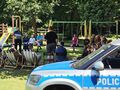 uczniowie siedzą na ławkach w parku i słuchają pani policjantki która stoi i mówi z przodu stoi radiowóz