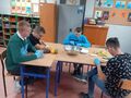 uczniowie siedzą przy ławce i kroją owoce na sałatkę
