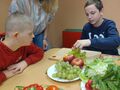 uczniowie siedzą przy stole gdzie znajduja sie owoce jeden z nich nakłada winogrono na gruszkę tworzac jeża