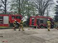 Strażacy biegną trzymając w ręku sprzęt gaśniczy. W tle wozy strażackie 