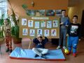 Troje dzieci stoi obok tablicy z rysunkami jedno dziecko siedzi na materacu przed tablicą wszyscy są w holu