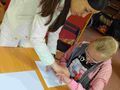 uczennica siedzi przy ławce w klasie i z pomocą nauczyciela klei obrazek