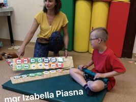 Pani Izabella Pisana znajduje się razem z uczniem w sali gimnastycznej . Nauczyciel prezentuje makietę z sylabami. Na dole zdjęcia napis: mgr Izabella Piasna