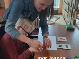 3. Pani Joanna Drobnikowska maluje razem z uczniem literę A. Używa do tego palca wskazującego dziecka i farb w kolorze czerwonym i niebieskim. Na dole zdjęcia napis: mgr Joanna Drobnikowska 
