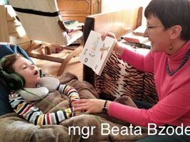 Uczeń siedzi w siedzisku z granulatu, ma założone słuchawki na uszach. Pani Beata Bzodek pokazuje książeczkę z samogłoskami. Na dole zdjęcia napis: mgr Beata Bzodek